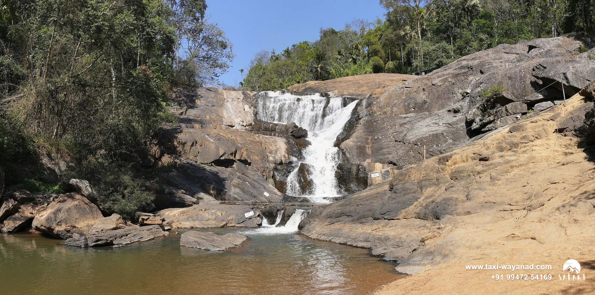 Calm pool and sandy bank below Kanthanpara Waterfalls framed by trees, Wayanad